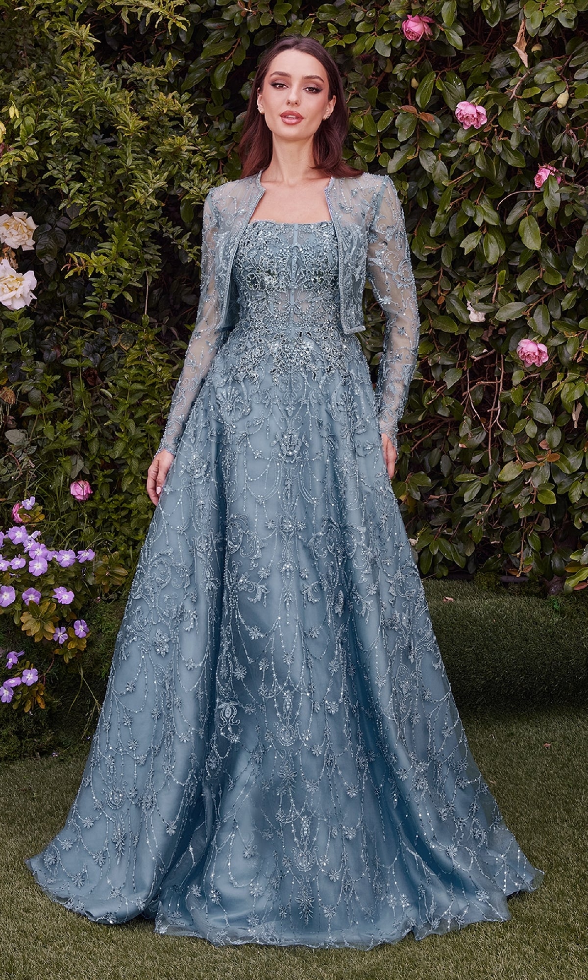 Amid lush greenery and blooms, a woman exudes elegance in the Andrea & Leo MA104: a light blue long sleeve formal gown featuring embroidered lace, intricate beading, and a matching jacket for a graceful evening look.